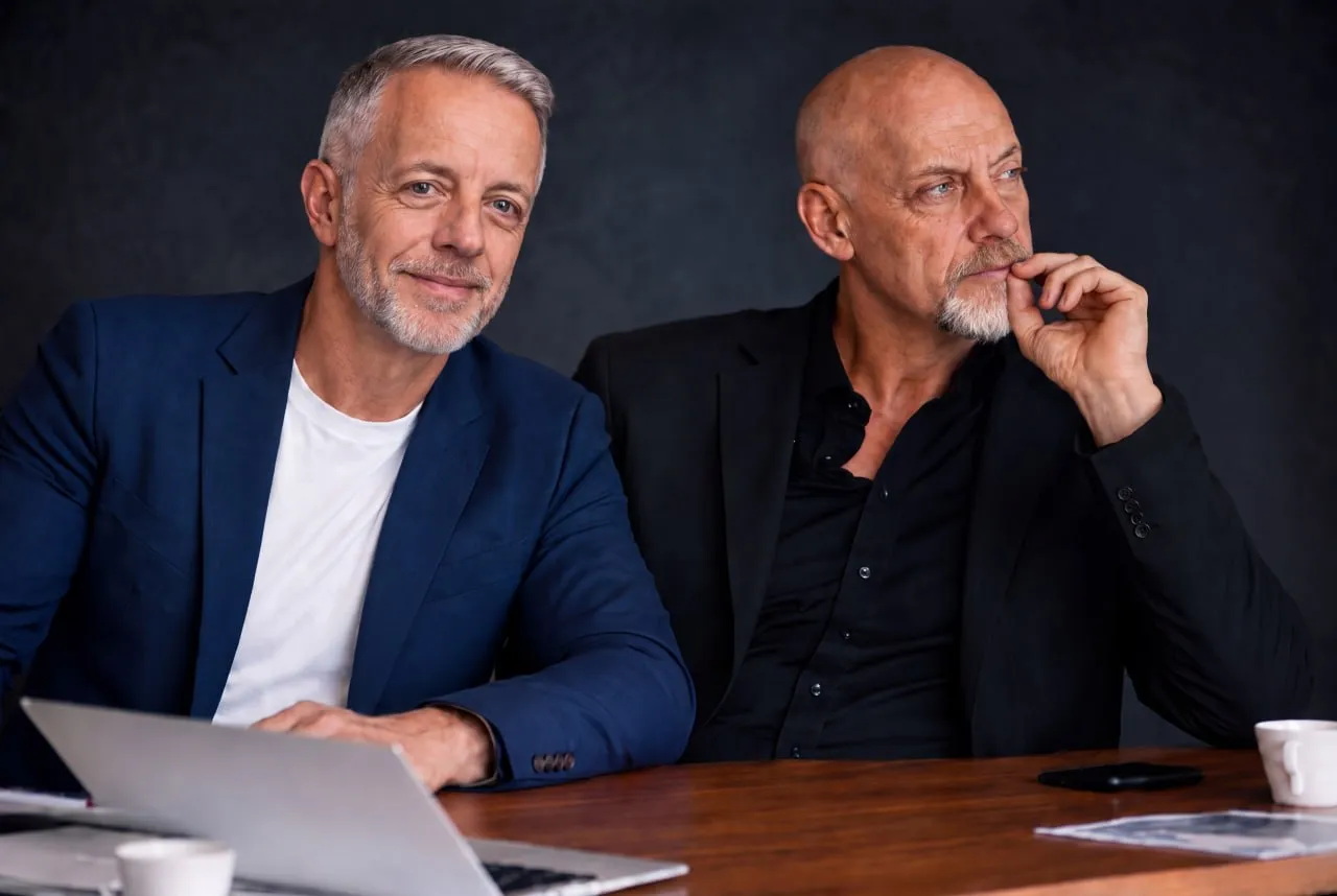 Damir Tomicic and Thomas Bothe sitting side by side at a wooden table, one looking into the camera, the other looking away in thought.